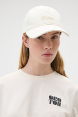 Profile view of model wearing the Oroton Oroton Script Cap in Antique White and 100% cotton canvas fabric for Women .